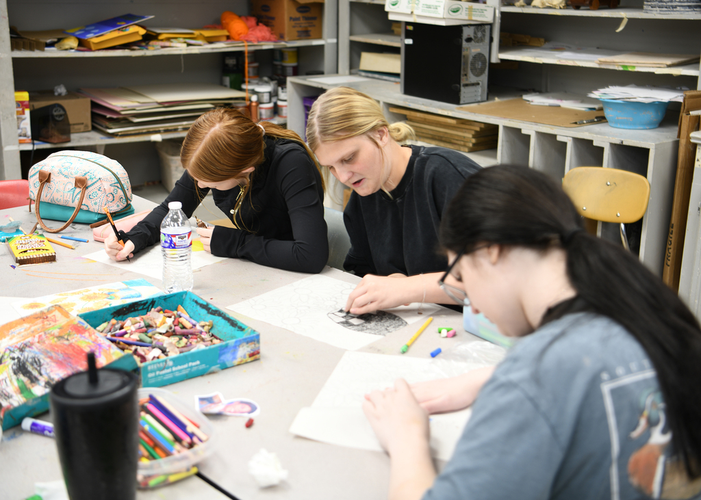 Three women sit at a table with art supplies. One writes, another draws, and the third looks on.