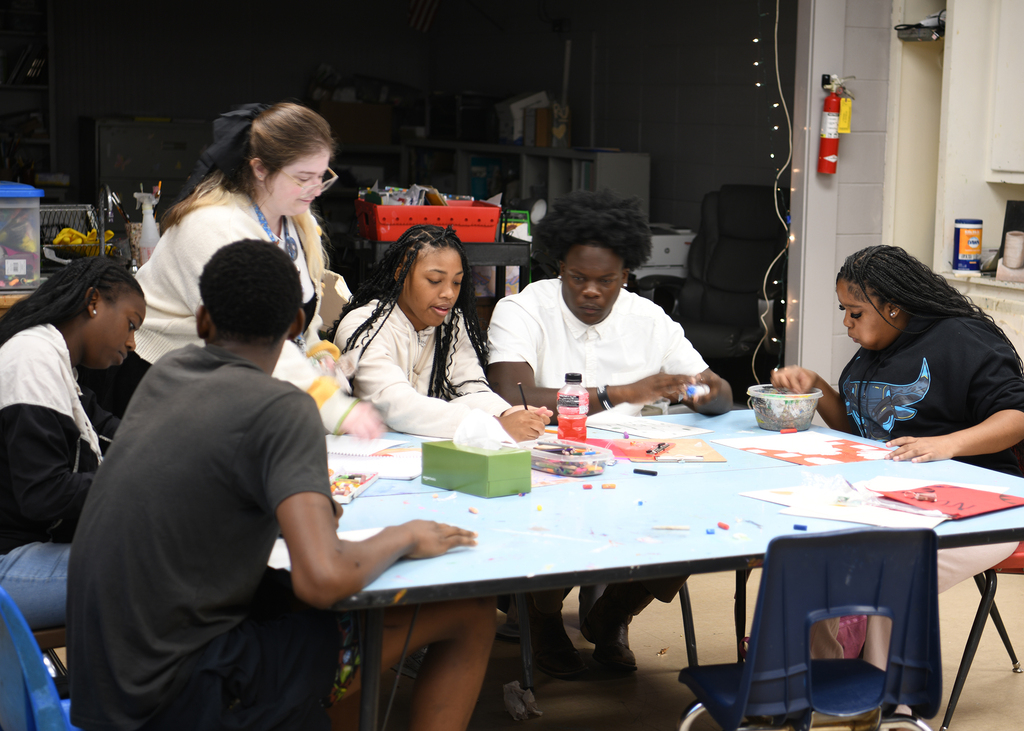 Students are sitting at a table, with art supplies and crafts, wearing casual clothing.