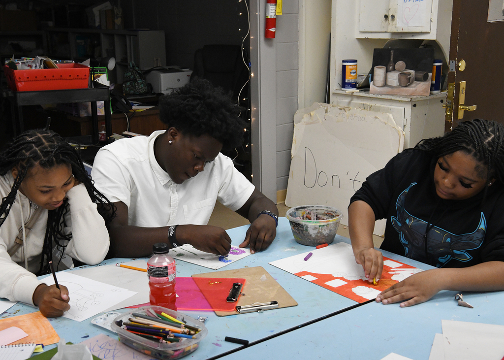 Three individuals work on a project at a table. One uses a marker, another holds a paper, and the third has a box of crayons.
