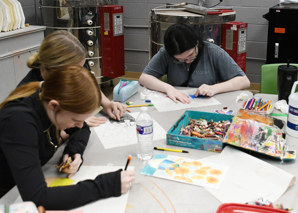 Three women create art at a table, surrounded by papers, colored pencils, and other materials.