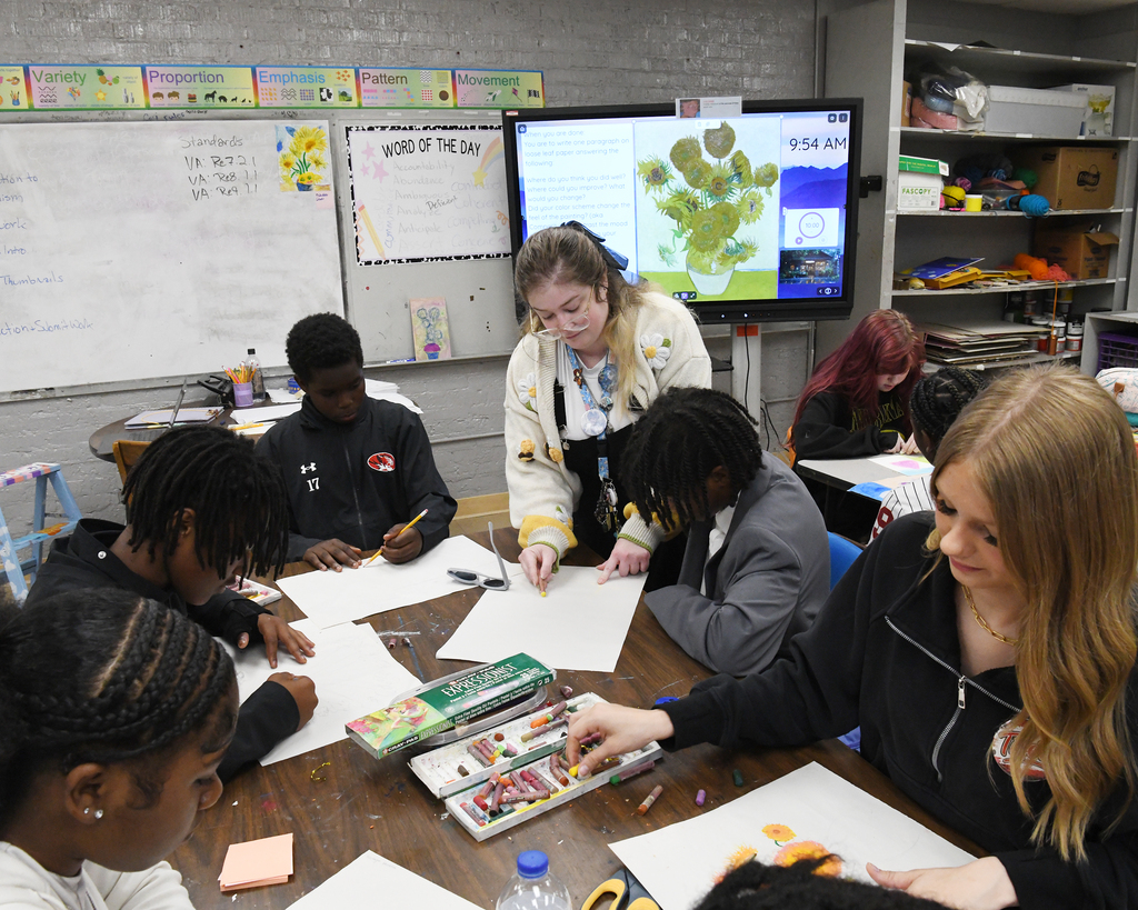 A classroom with several students sitting at a table. Two adults are helping them. A monitor displays a colorful image.