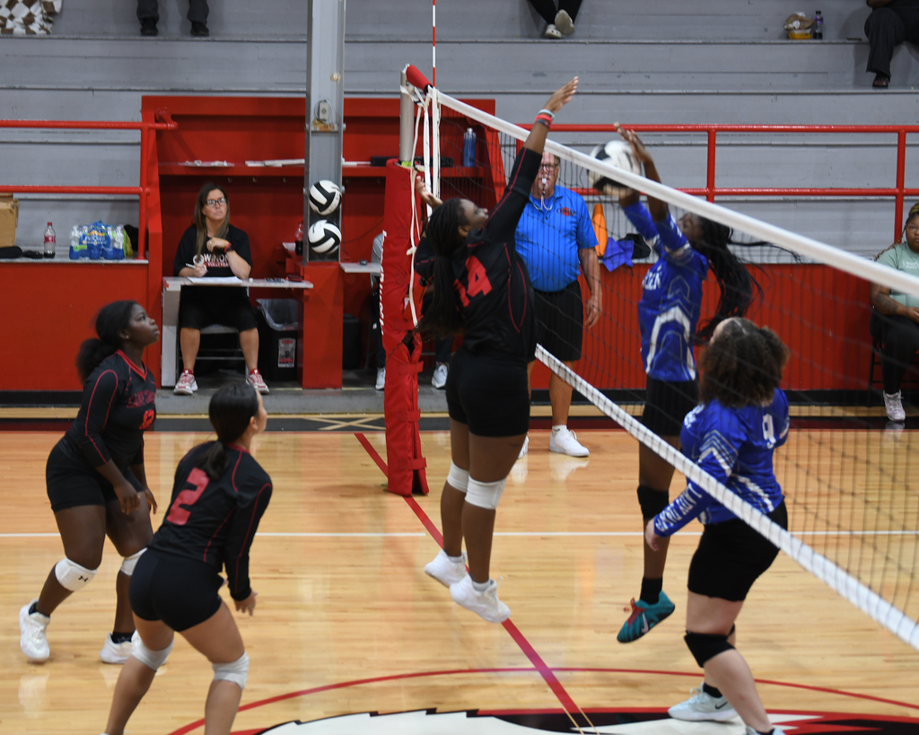 Players in matching uniforms on two teams. One team in black, the other in blue. They play volleyball.