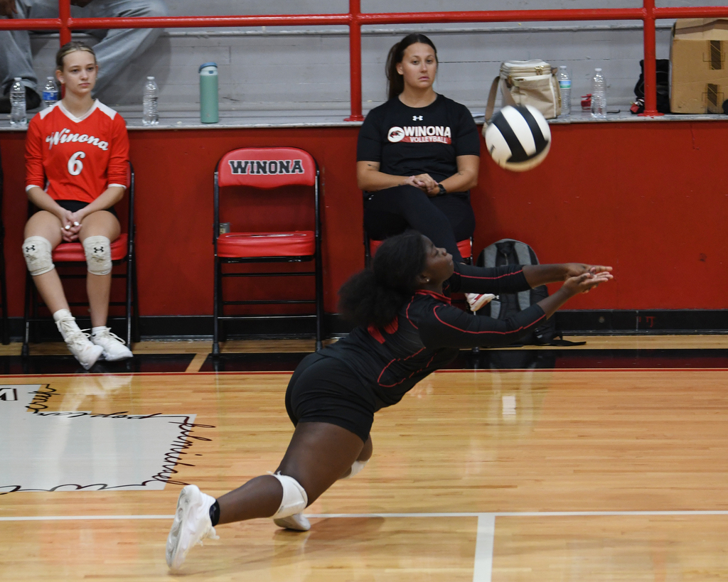Player in black diving on a wooden court, two teammates in red and black jerseys seated at a red wall.