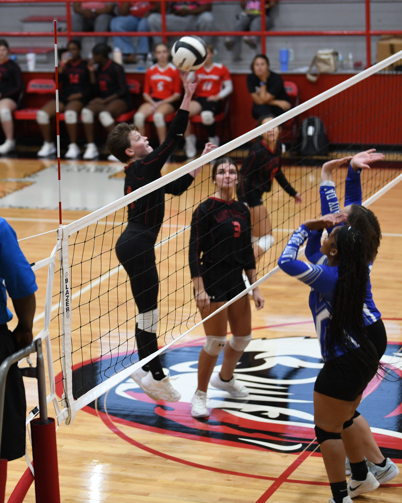 Four female volleyball players on an indoor court. Two players on each side of a net.