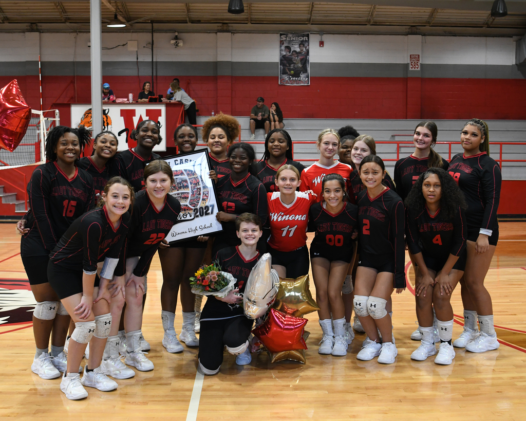 A sports team poses for a group photo on a court with a red and white wall in the background.