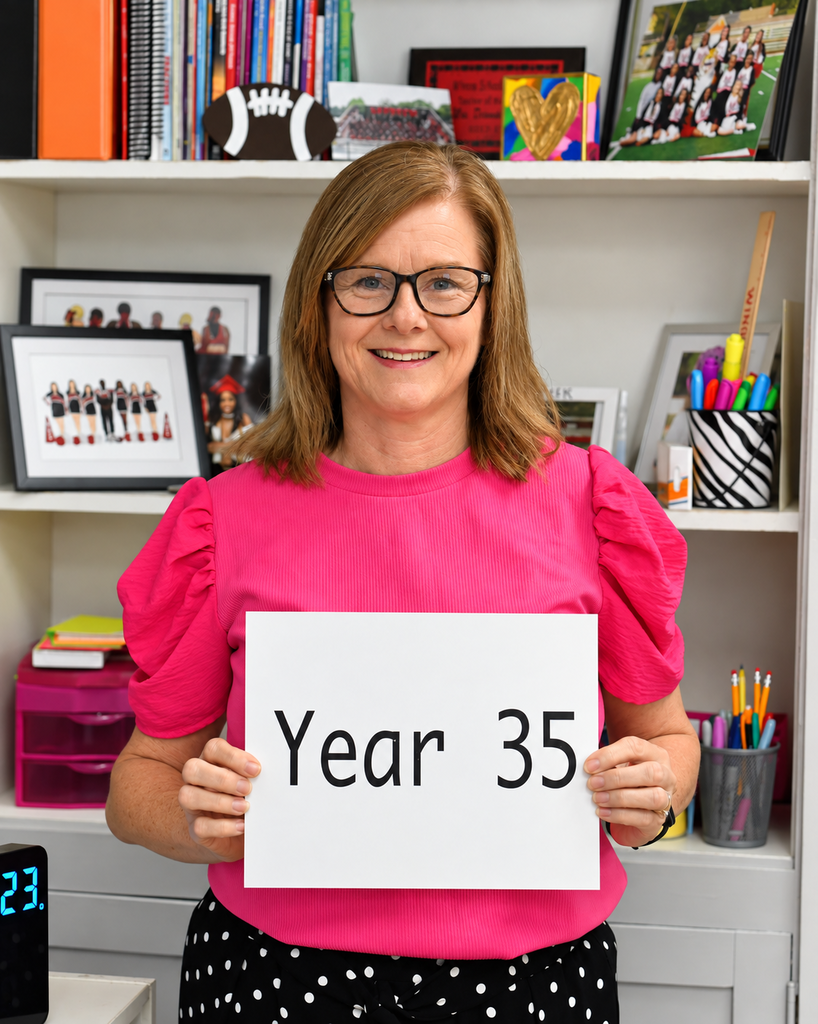A woman in a pink blouse holds a sign reading "Year 35." Behind her, a bookshelf holds books, framed pictures, and office supplies.
