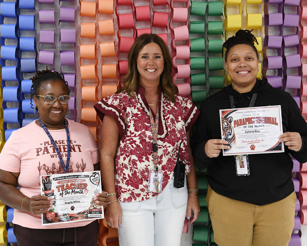 Three women standing behind a colorful backdrop. Each holds a certificate. One has a pink shirt, one a red floral blouse, and the last a black hoodie.