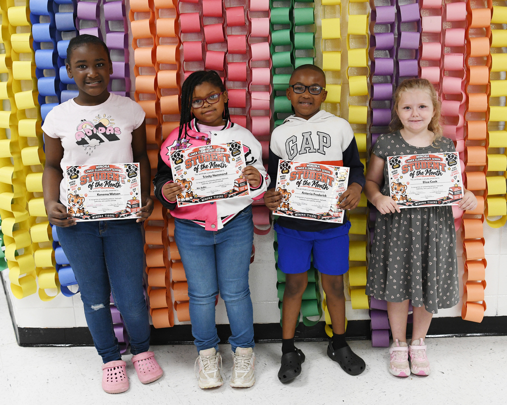 Four children hold certificates in front of a wall decorated with colorful paper chains.