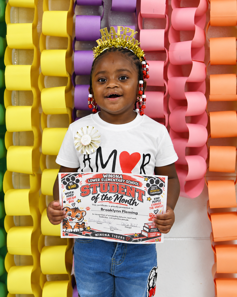 A child holds a certificate for student of the month, smiling. They stand in front of colorful paper chains.