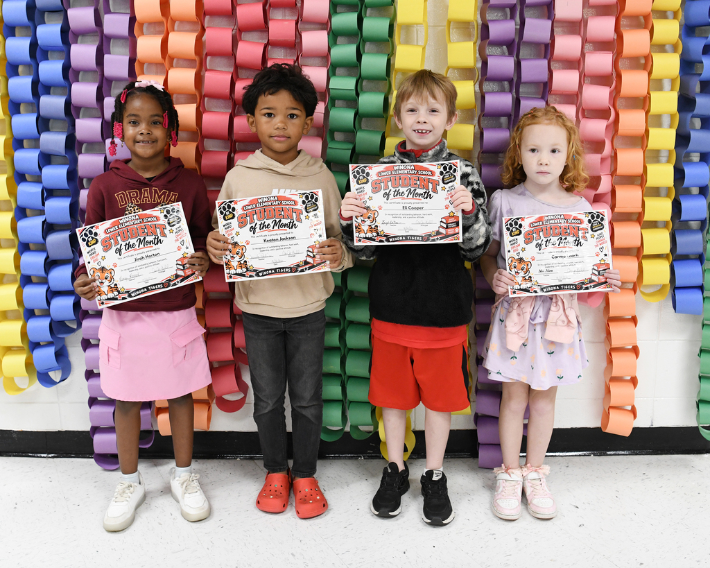 Four children stand in a row holding certificates, smiling at the camera against a backdrop of colorful paper chains.