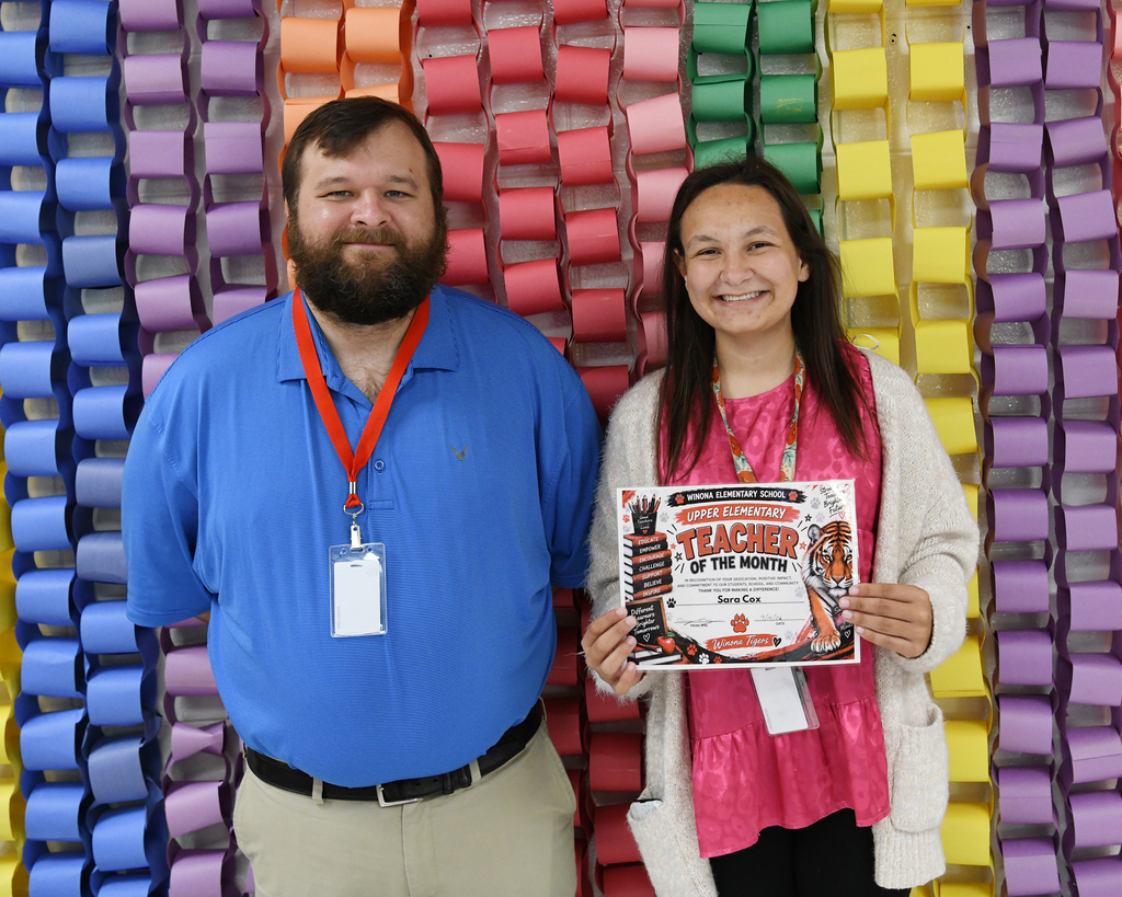A man and woman stand in front of a colorful background, smiling. The woman holds a certificate.