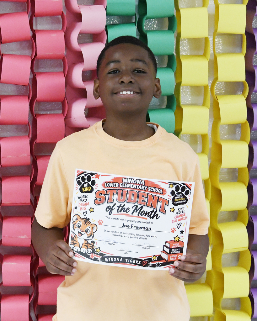 A smiling young boy holds a certificate reading "Student of the Month" in front of a wall of multicolored paper links.