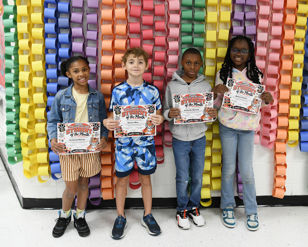 Four children stand in a row holding certificates, posing against a backdrop of colorful paper loops.