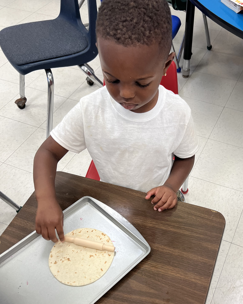 Child seated at a table, using a rolling pin to flatten a tortilla on a silver tray.