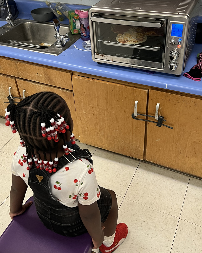 A young girl sits on a stool in a kitchen, looking into an oven. Kitchen counter with sink and cabinets behind.