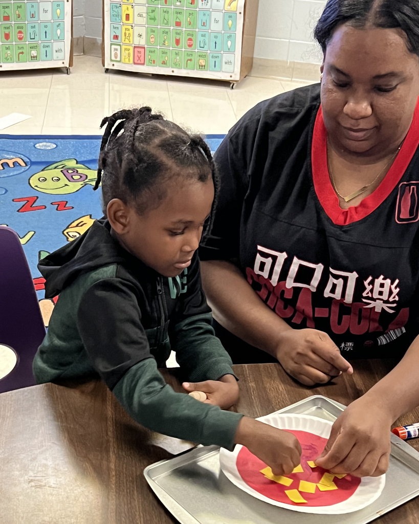 A woman helps a child with a craft project at a table, surrounded by classroom items.