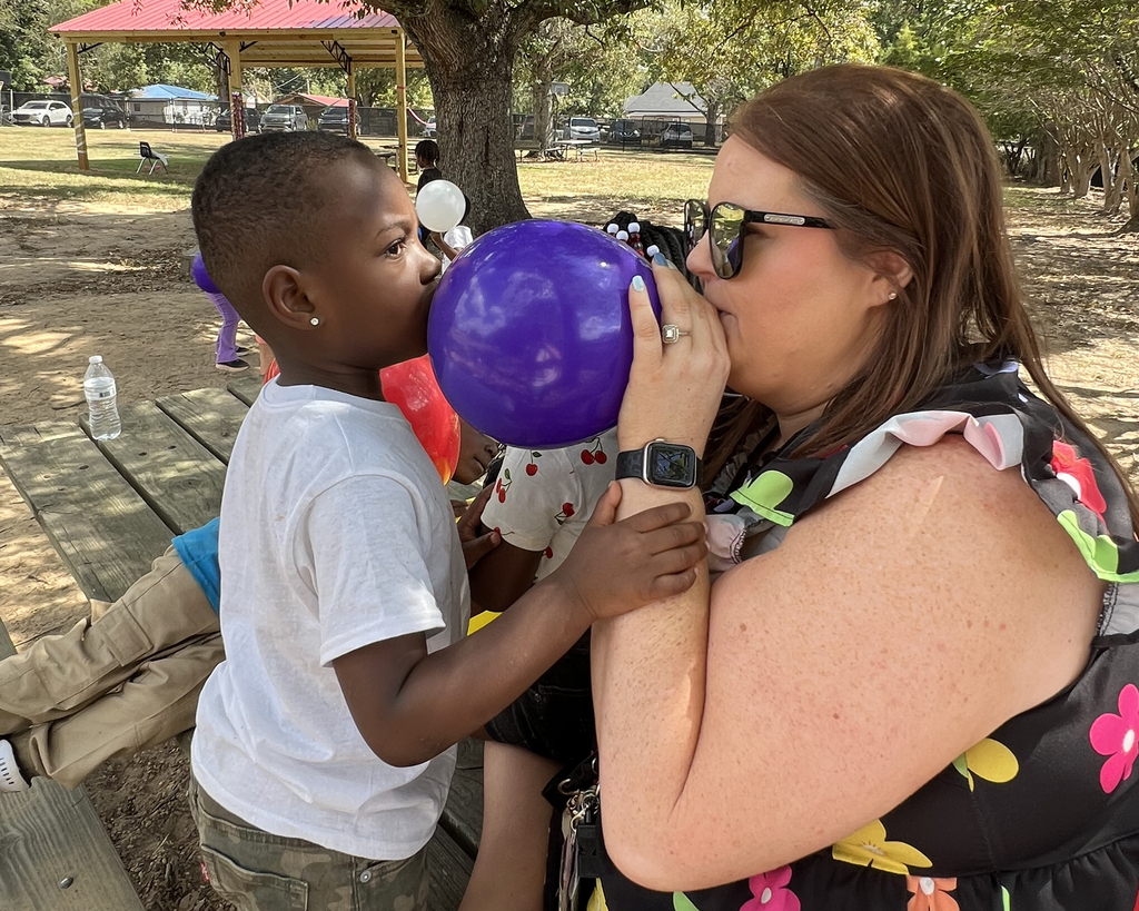 A woman blows up a purple balloon while a young child watches, seated on a bench in a park.