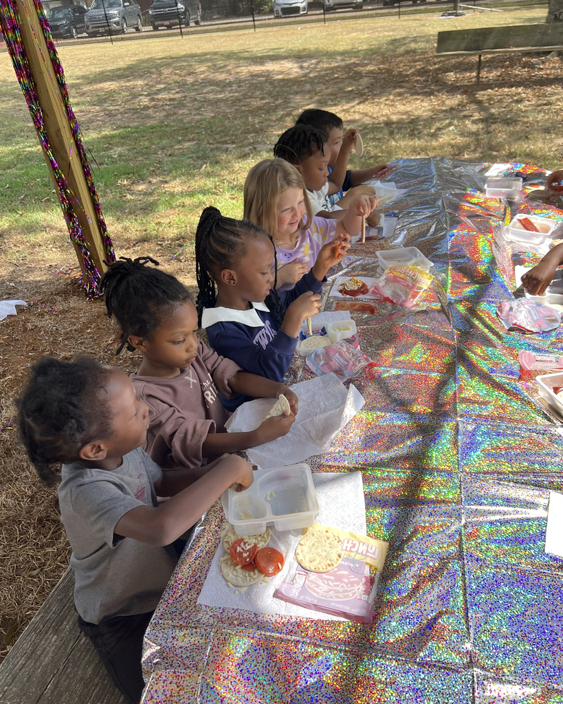 Kids seated around a long table outdoors, preparing meals with utensils and food in front of them.