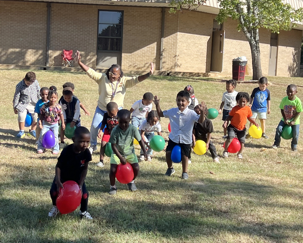 A group of children holding balloons runs outside near a building. Some balloons are red, yellow, and blue.