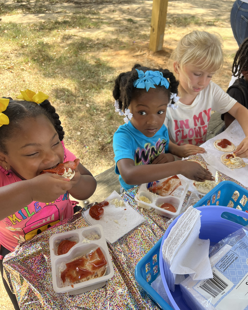 Three children seated at a picnic table, eating pizza and other foods. One child wears a yellow bow.