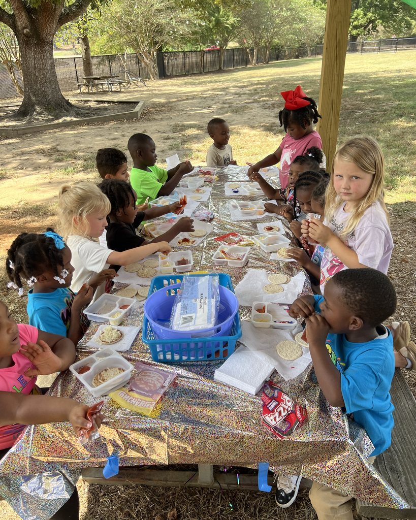 Children sit around a table outdoors, eating. Some wear white, others wear blue, and a few have headbands.