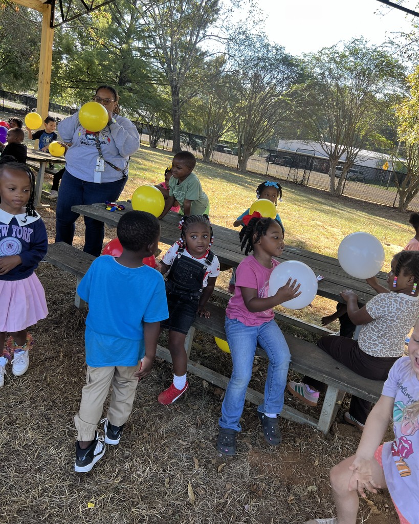 Several children, some holding balloons, sit and stand around a picnic table in a park. Trees and grass in the background.
