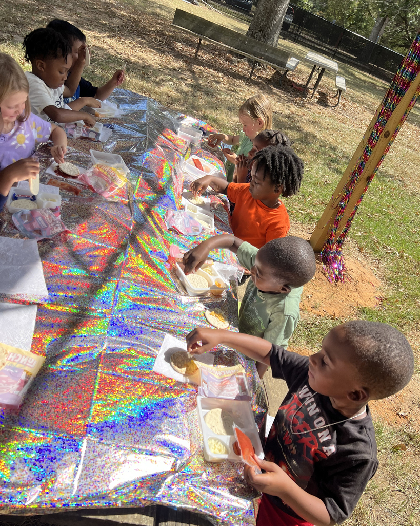 Children eating at a table with a multicolored tablecloth. A grassy area is in the background.