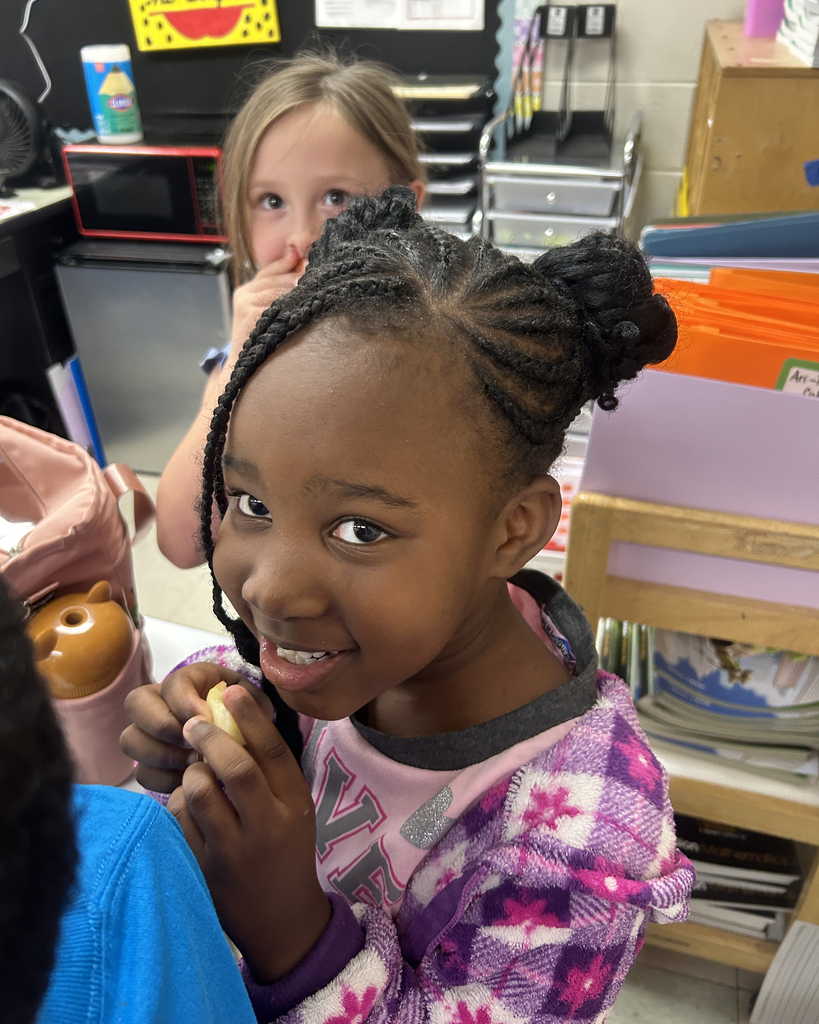 A young girl with braided hair smiles while holding food in a classroom setting. Another child is visible in the background.