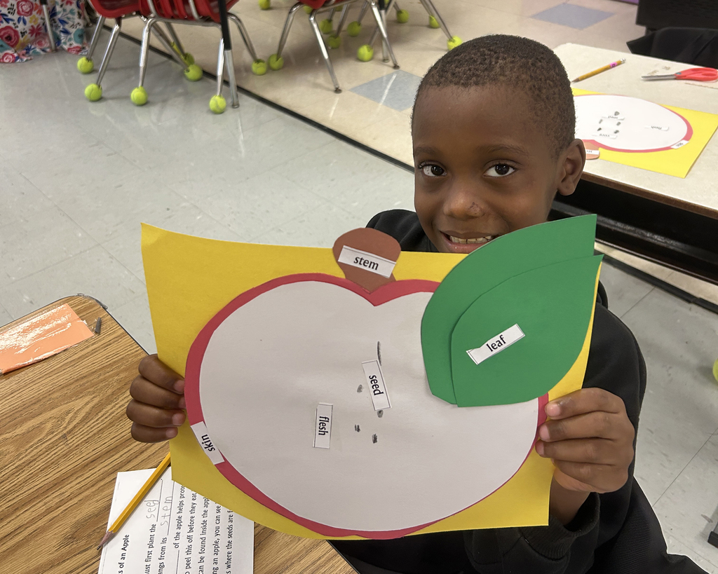 A child holds up a paper apple with a green leaf attached, smiling. Behind them is a classroom setting with chairs and tables.