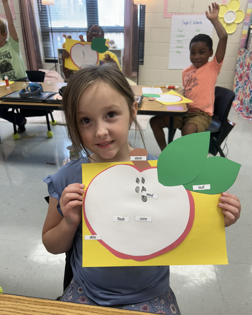 A young girl holds an apple-shaped paper craft with a smiling face and leaves, sitting in a classroom.