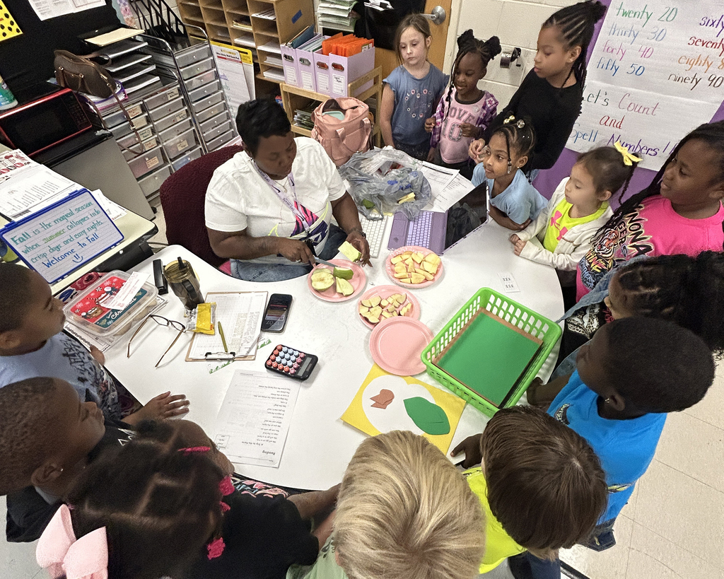 An adult leads a group of young children in a classroom. Children sit around a table with food, learning materials, and a calculator.
