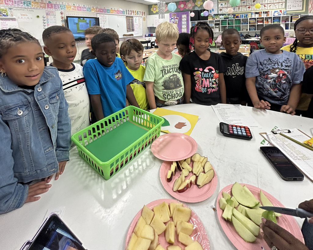 A classroom setting with several young children around a table with food. Tables have plates of cut apples and utensils.