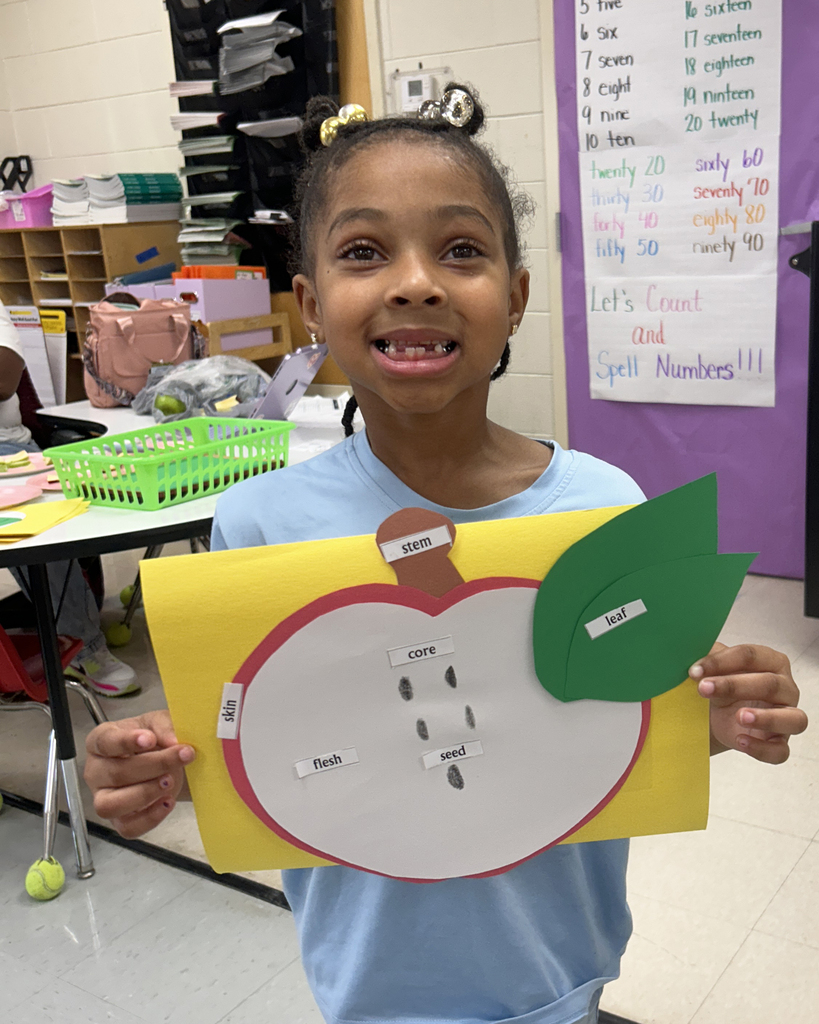 Child in a classroom holds an apple-shaped cutout with a number pattern and leaf. Behind, a classroom with tables and storage boxes.
