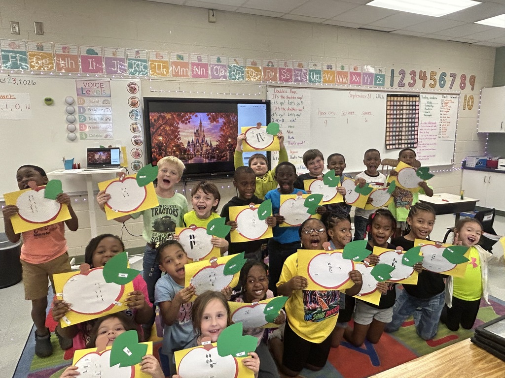 A classroom with children in yellow shirts holding up paper apples and smiling at the camera.