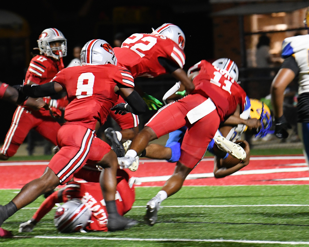 Football players in red and blue uniforms collide on a field. Players wear helmets and gloves. Background shows a blurred view of a building and people.