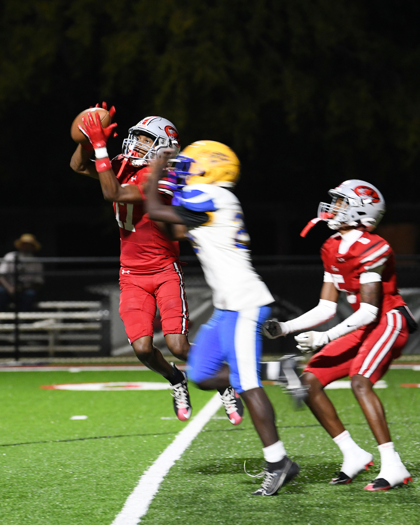 Three football players on a field. Two wearing red, one in blue and white. The player in red catches a ball.
