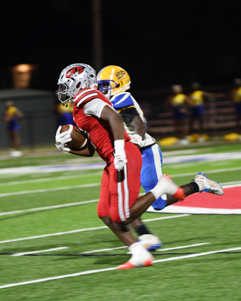 Two football players wearing helmets and uniforms run on a field. One player holds a football while the other pursues.