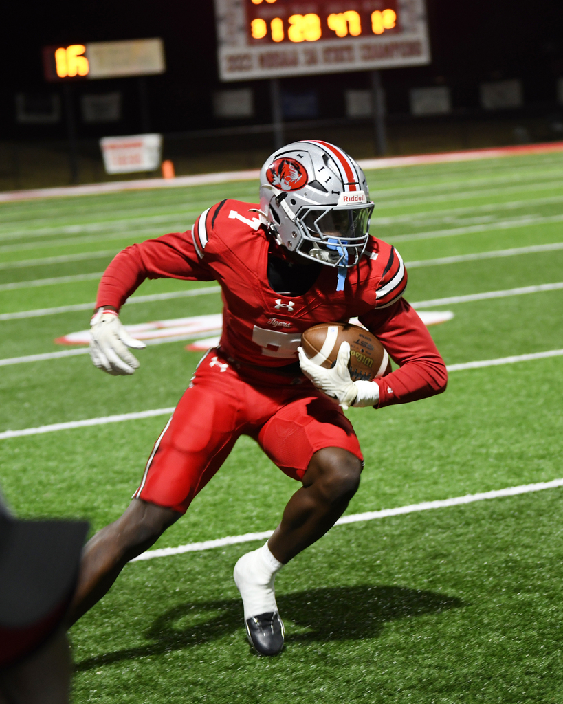 A football player in red gear and helmet running on a field with a scoreboard in the background.