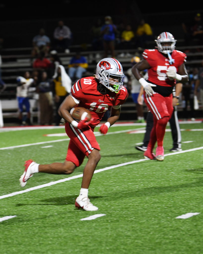 A player in red runs with a football on a field. Behind him, another player in red and white runs. Audience members watch from the stands.