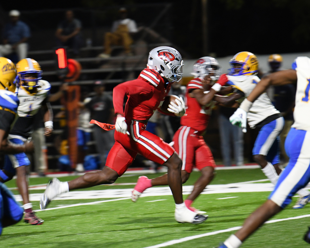 Players wearing helmets and jerseys, running on a field. A player in red runs with a ball.