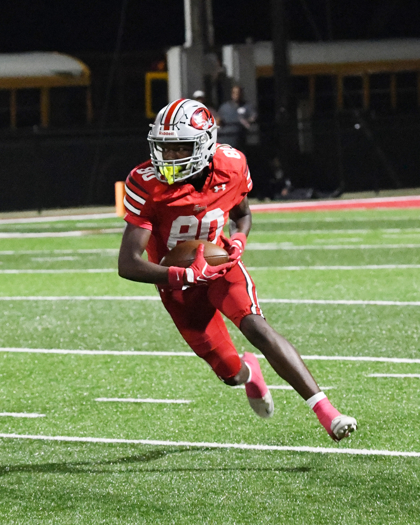 Athlete in red uniform, helmet, gloves, and pink socks jumps in air with football during football game.