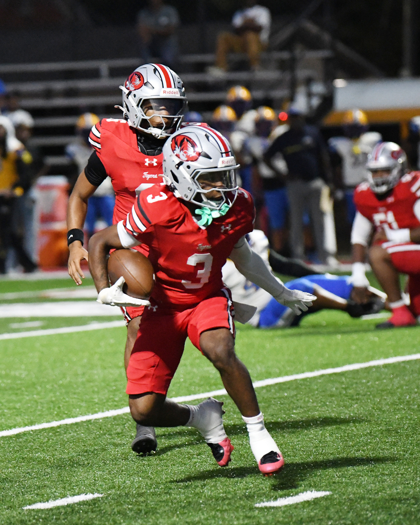 Two football players in red uniforms with one holding a ball, running on a field with white lines.