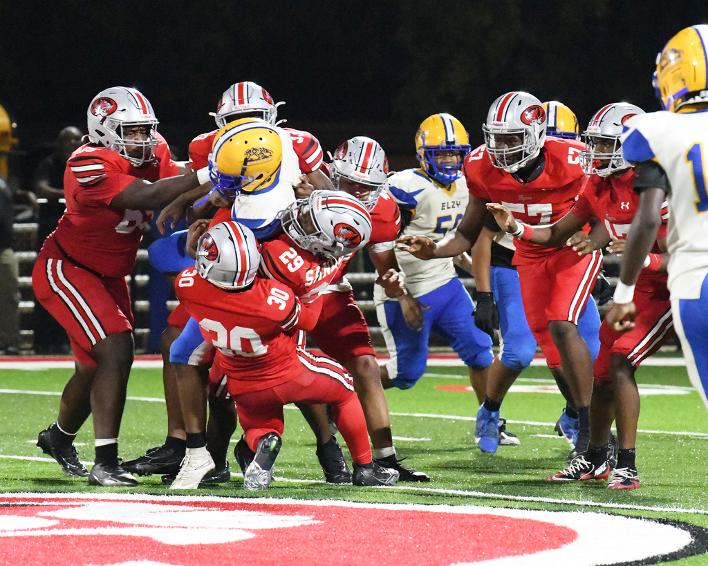 Football players on a field engage in play. They wear various colored uniforms and helmets. The ground features a red-and-white circular pattern.