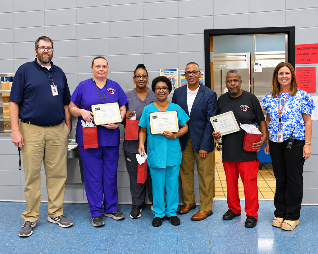 A group of six people in a room holding certificates, one woman in blue scrubs stands out.