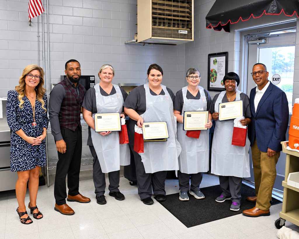 Five adults in aprons hold certificates, standing with four others in a room. Behind them, a door leads to outside.