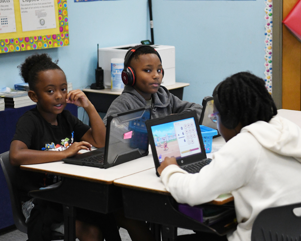 Three students are seated at a table using laptops. One has headphones on. Behind them is a bulletin board.
