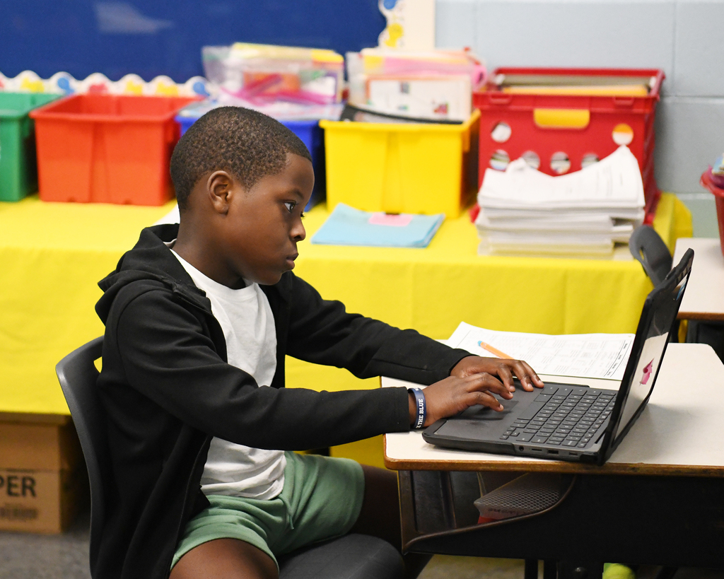 A young boy sits in a chair at a desk, typing on a laptop, with storage bins and a yellow tablecloth in the background.