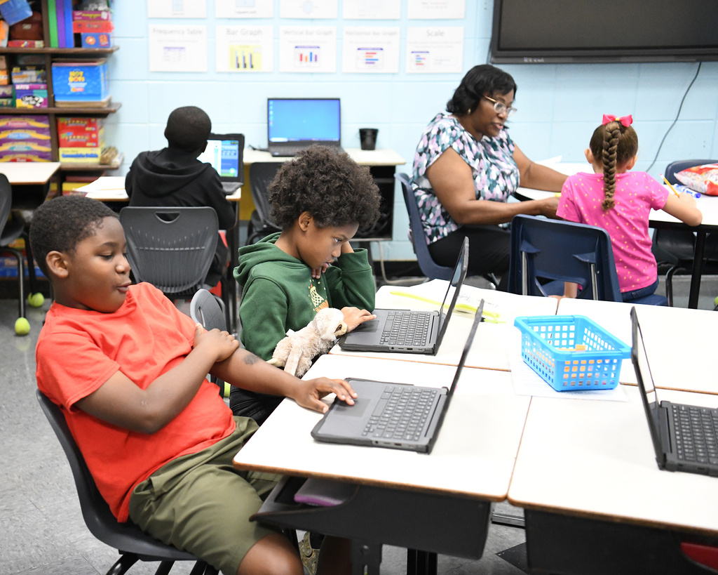 Four children and two adults in a classroom, with laptops on tables and a monitor on the wall.