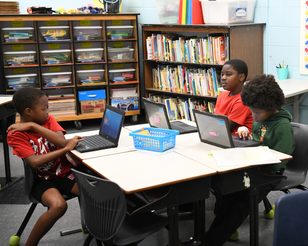 Four children sit at a table with laptops, against a backdrop of shelves filled with books.