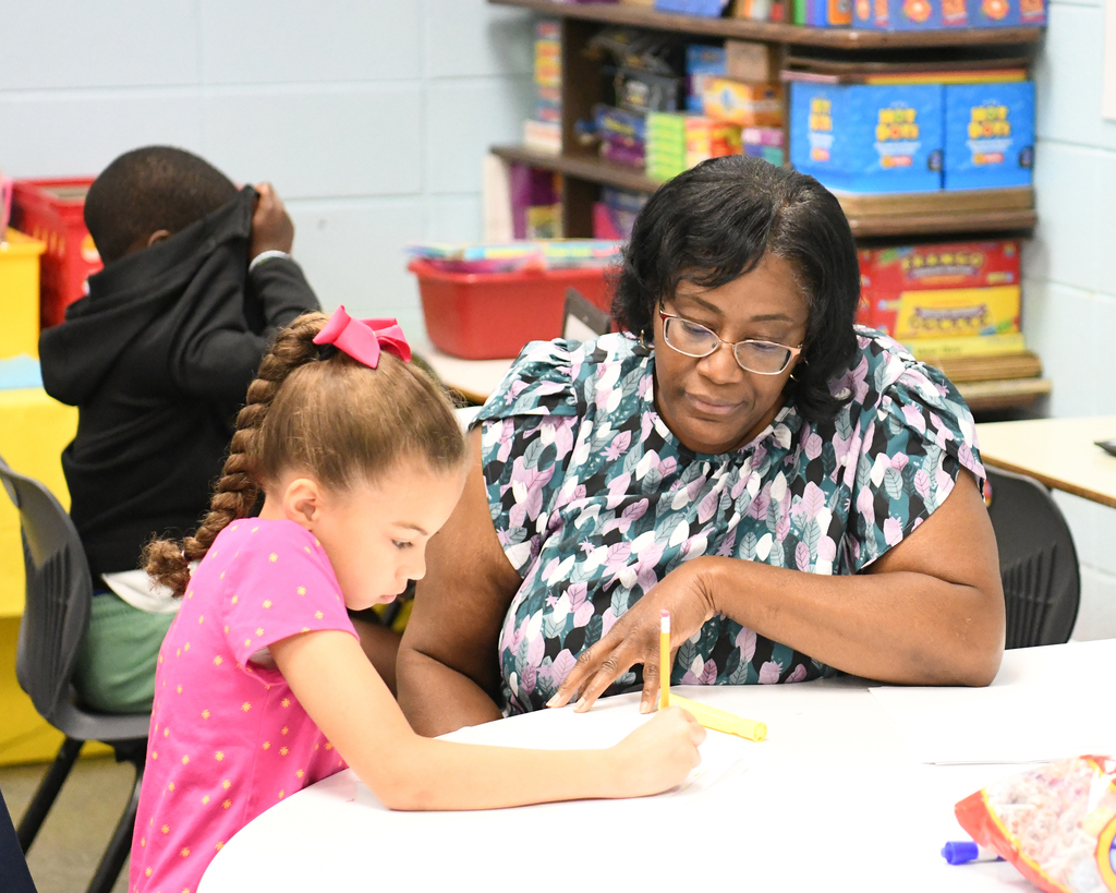 A woman and a girl in a classroom, seated at a table, both holding pencils. Shelves and storage boxes in the background.